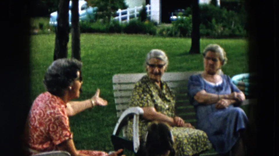 The home-movie frame shows a red-dressed woman addressing two elderly women on a bench in a suburban Hicksville yard.