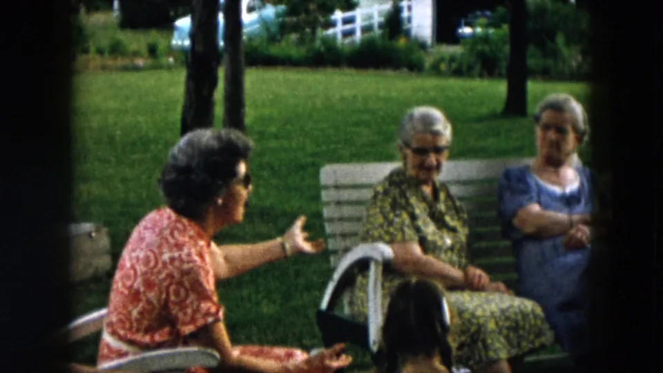 Three elderly women remain gathered around a white bench on a green Hicksville backyard lawn, with trees and fencing beyond.