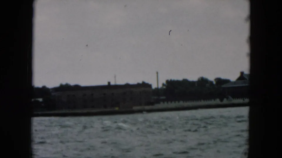 A soft 8mm view of low waterfront structures, trees, and a smokestack across choppy water in the supplied 1957 New York clip.