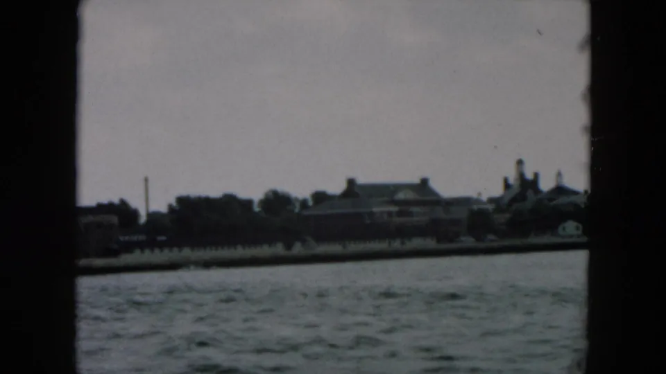 1957 8mm frame from a moving vessel showing distant shoreline buildings across gray water in the Hicksville, New York metadata context.