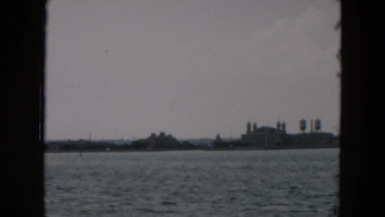1957 8mm view across New York Harbor toward a hazy shoreline with industrial-looking structures and water towers.