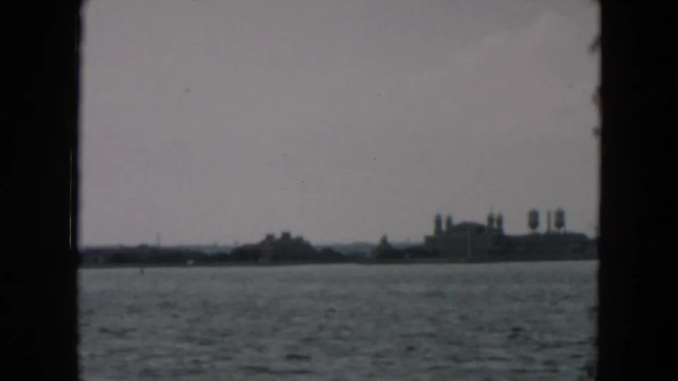 1957 8mm view across New York Harbor toward a hazy shoreline with industrial-looking structures and water towers.