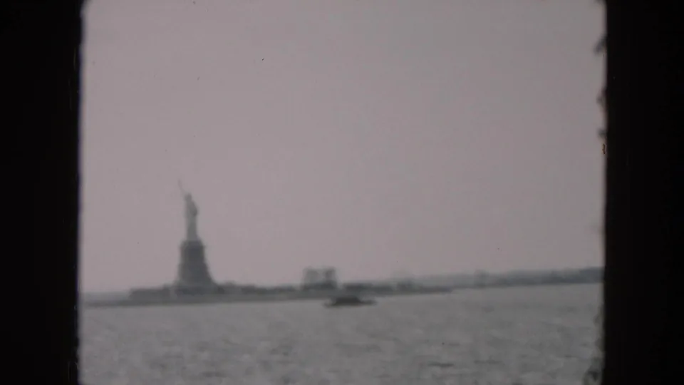 Distant 1957 8mm harbor view of the Statue of Liberty on Liberty Island, with a small watercraft crossing the foreground.