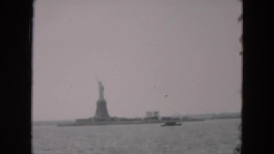 Soft 8mm frame of the Statue of Liberty from New York Harbor in 1957, with Liberty Island and a small boat visible through haze.