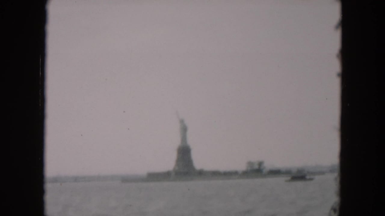 1957 8mm home-movie view of the Statue of Liberty from across the water, with a hazy sky and dark border around the frame.