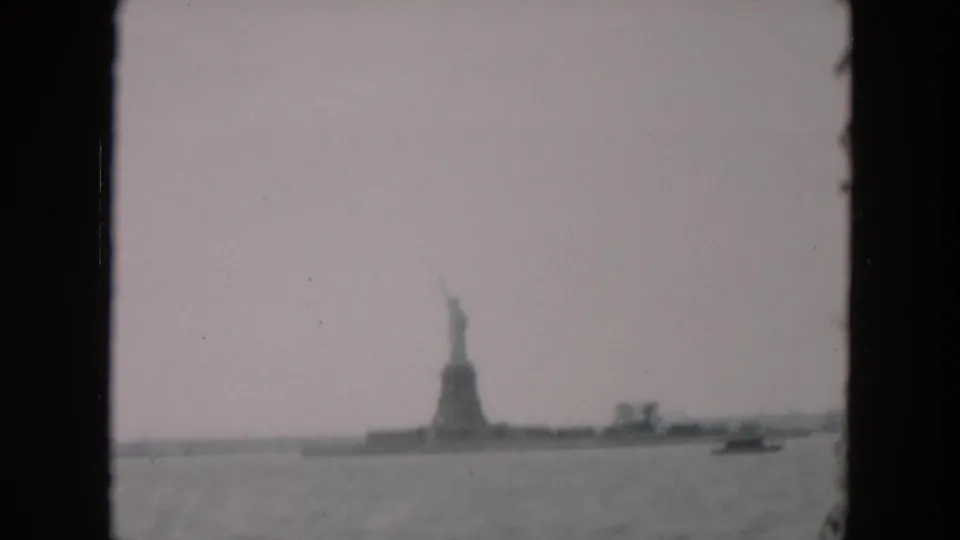 1957 8mm home-movie view of the Statue of Liberty from across the water, with a hazy sky and dark border around the frame.