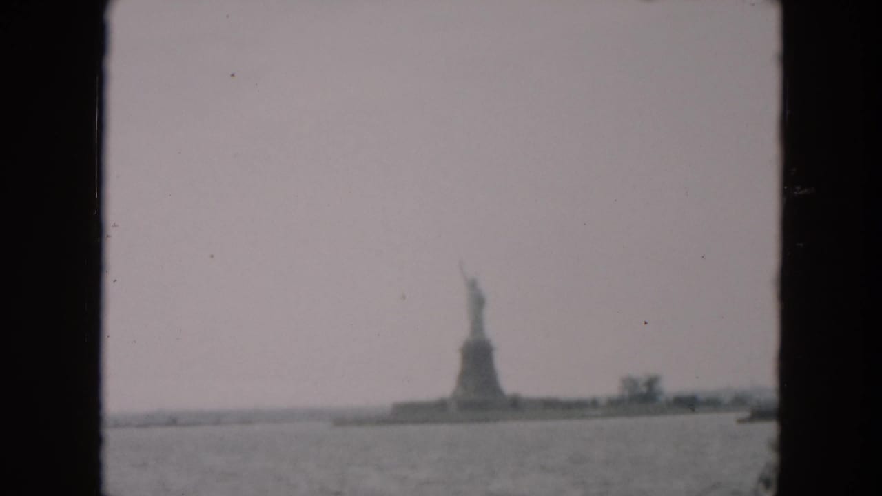 Distant 1957 home-movie view of the Statue of Liberty and Liberty Island, with harbor water and a small boat visible to the right.