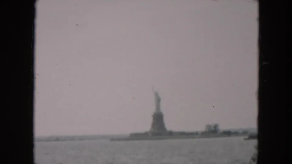 Distant 1957 home-movie view of the Statue of Liberty and Liberty Island, with harbor water and a small boat visible to the right.