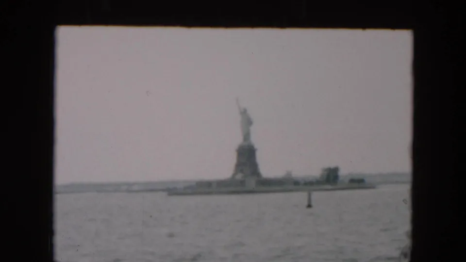 Distant 1957 8mm view of the Statue of Liberty and island shoreline, with muted color, grain, and harbor water in front.