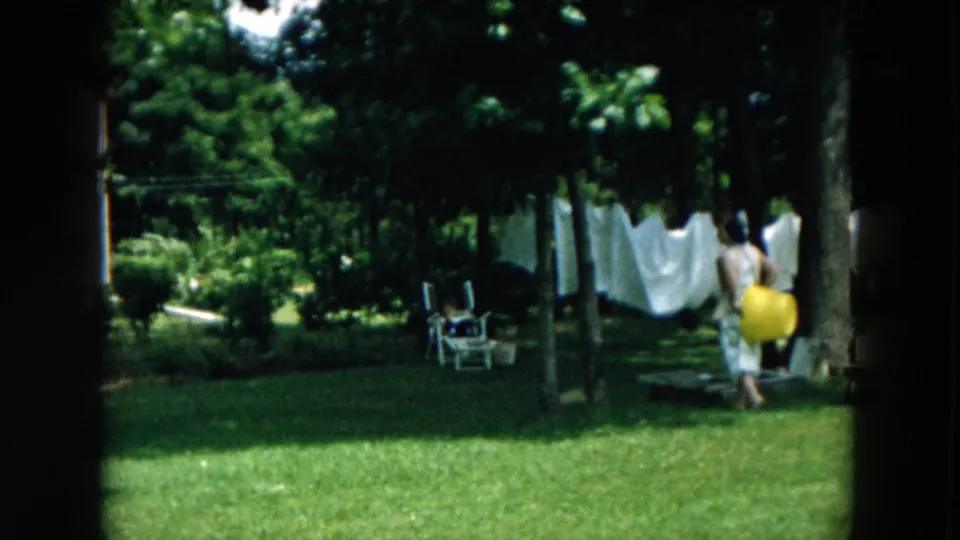 In a Hicksville backyard, a woman in a light dress walks past a clothesline with white laundry while holding a yellow bucket.