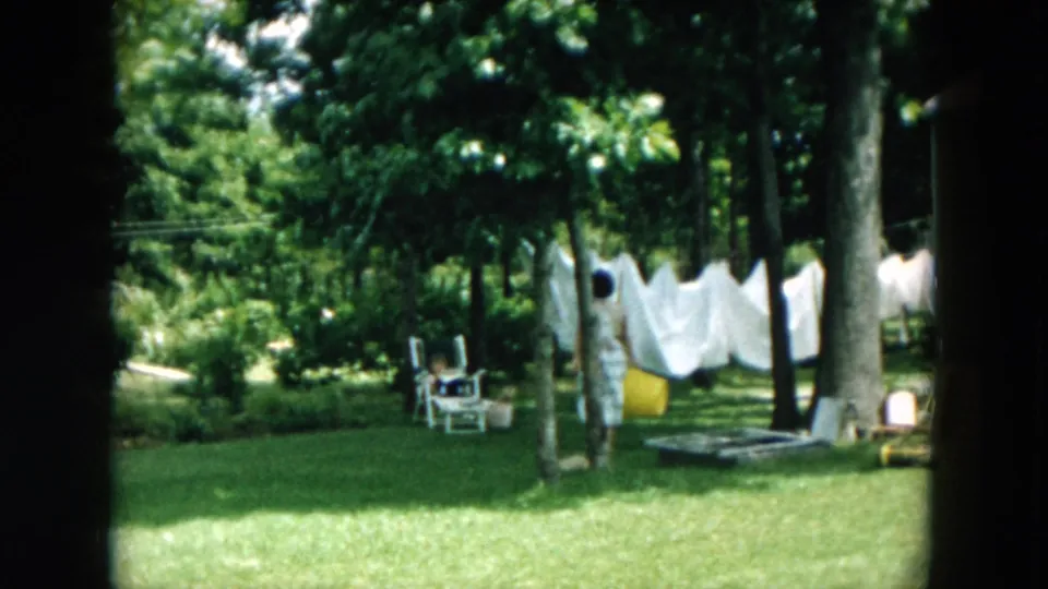 The woman appears near a backyard clothesline, with white laundry, a yellow bucket, chairs, trees, and grass visible around her.