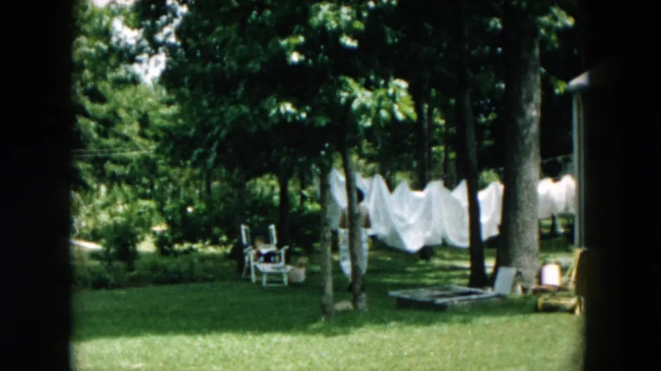 A 1957 Hicksville backyard frame shows white laundry on the line, lawn chairs, trees, grass, and the woman partly visible near the left edge.