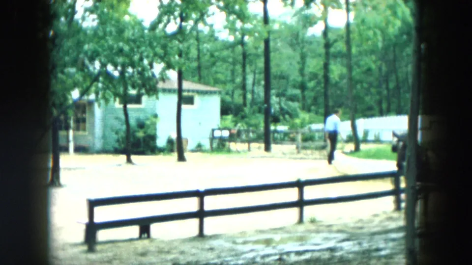 1957 Hicksville home-movie frame showing a wooded roadside, small light buildings, fencing, and a distant person walking.