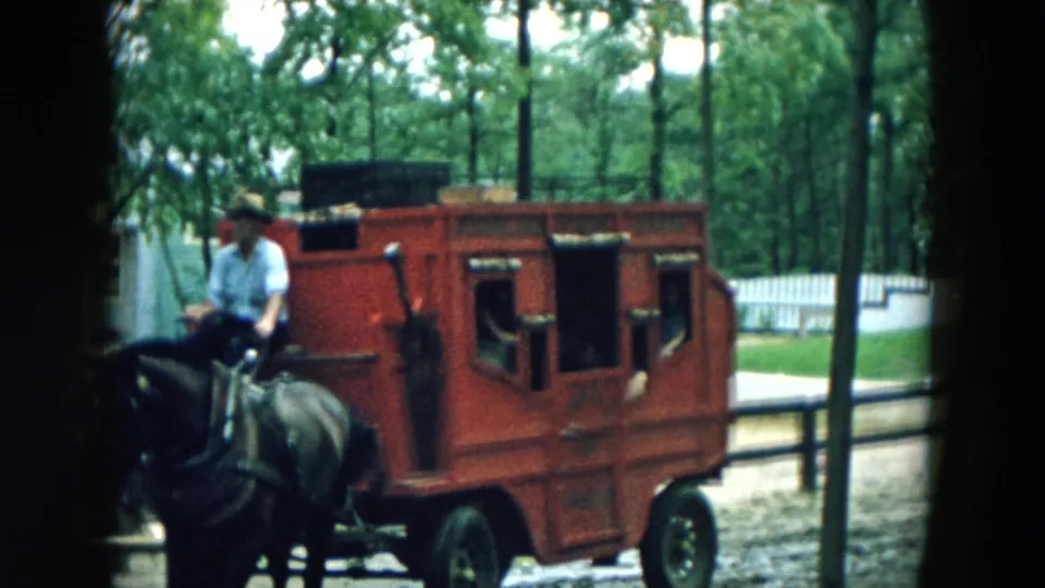 The red stagecoach continues past the camera, driven by a man in a light shirt and hat in the wooded Hicksville scene.
