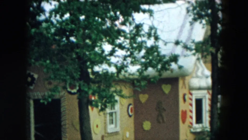 1957 Hicksville home-movie frame showing a storybook gingerbread-style playground house behind trees, with hearts and candy-like trim visible.