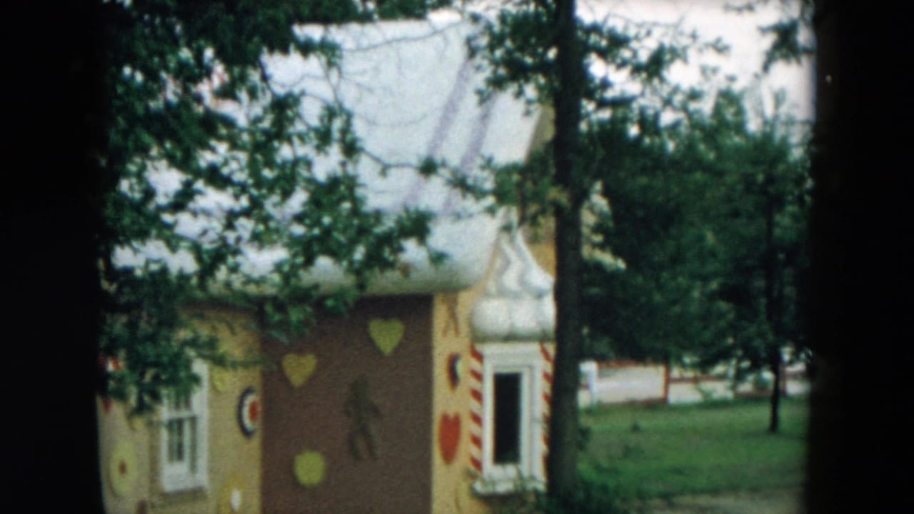 1957 Hicksville frame of a whimsical playground house with a white roof, painted hearts, candy-striped trim, trees, and open lawn.