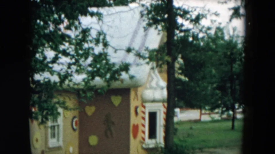 1957 Hicksville frame of a whimsical playground house with a white roof, painted hearts, candy-striped trim, trees, and open lawn.