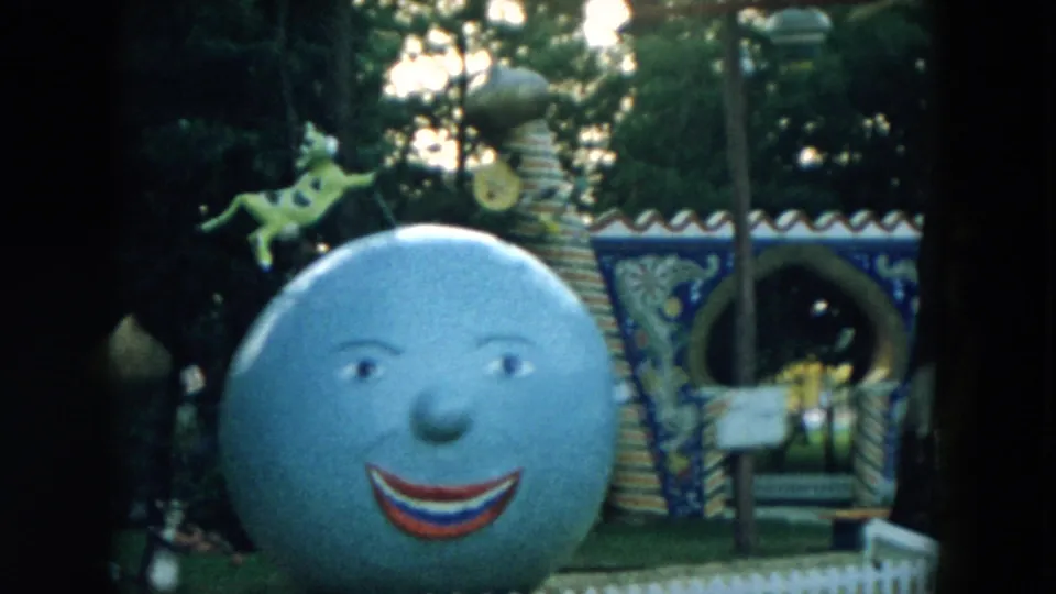 1957 Hicksville home-movie frame showing a large smiling blue moon sculpture near a painted arch and striped columns.