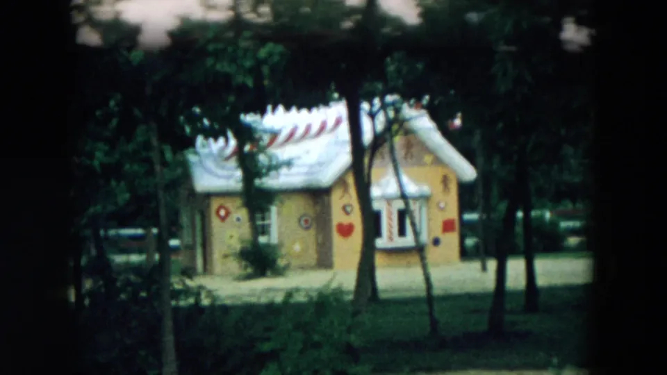 1957 Hicksville home-movie frame showing a whimsical painted playhouse with a white roof, hearts, circles, and trees in front.