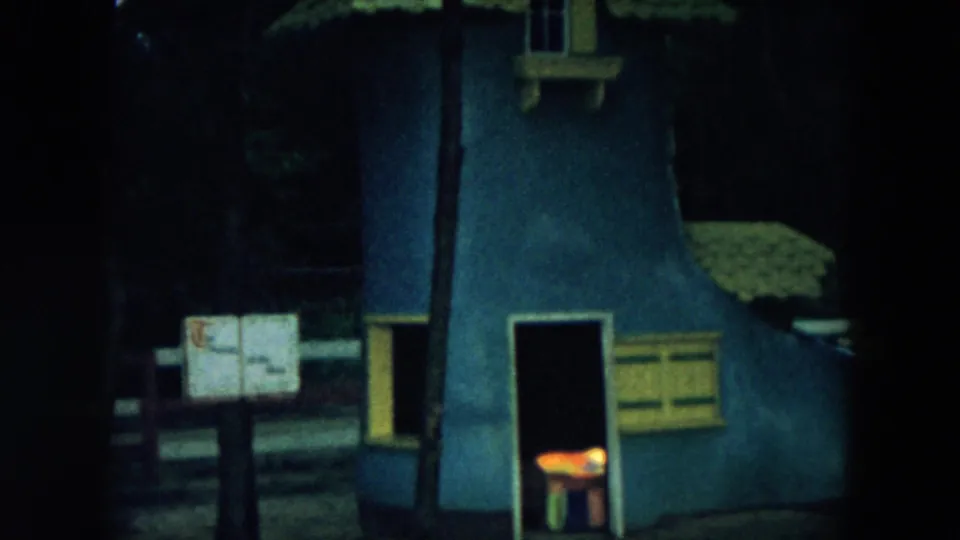 1957 Hicksville home-movie frame showing a blue boot-shaped novelty playhouse in a wooded setting, with a small lit opening and trees around it.