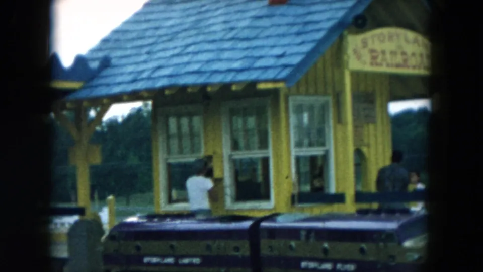 Hicksville Storyland Railroad station with visitors at the windows and a purple and gold train car crossing the foreground.