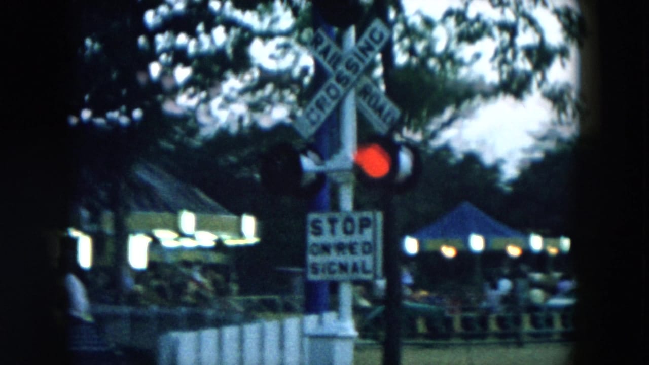 Railroad crossing signal with red light near shaded park structures.