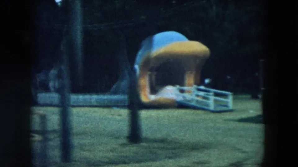 The moving train view passes a large dinosaur figure, fencing, poles, and a shaded walkway in the 1957 Storyland setting.