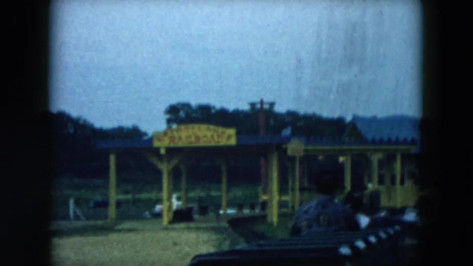 1957 Hicksville amusement-park ride view showing dark seats, a yellow station canopy, a sign, trees, and low buildings in soft 8mm color.