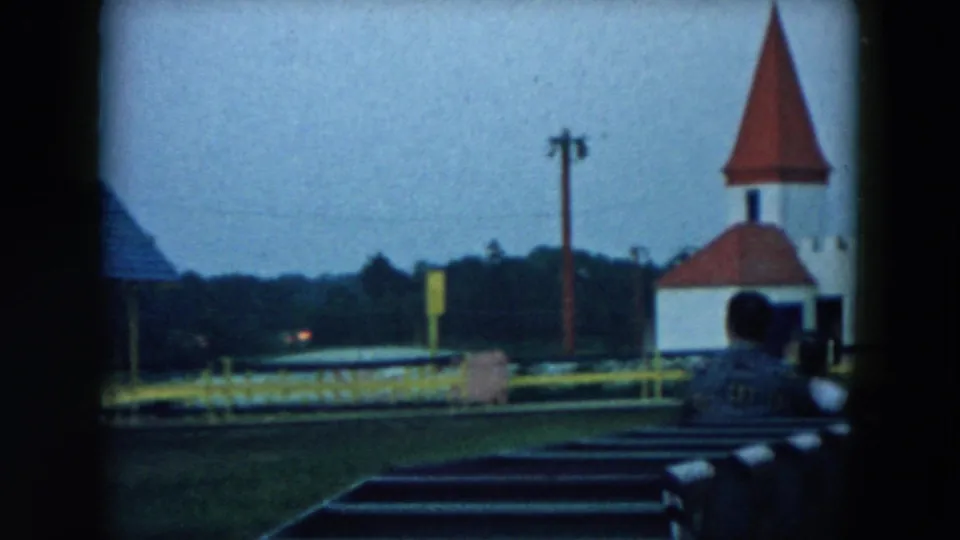 1957 Hicksville ride POV showing dark seats, a seated rider, yellow railing, and a small white building with a red conical roof.