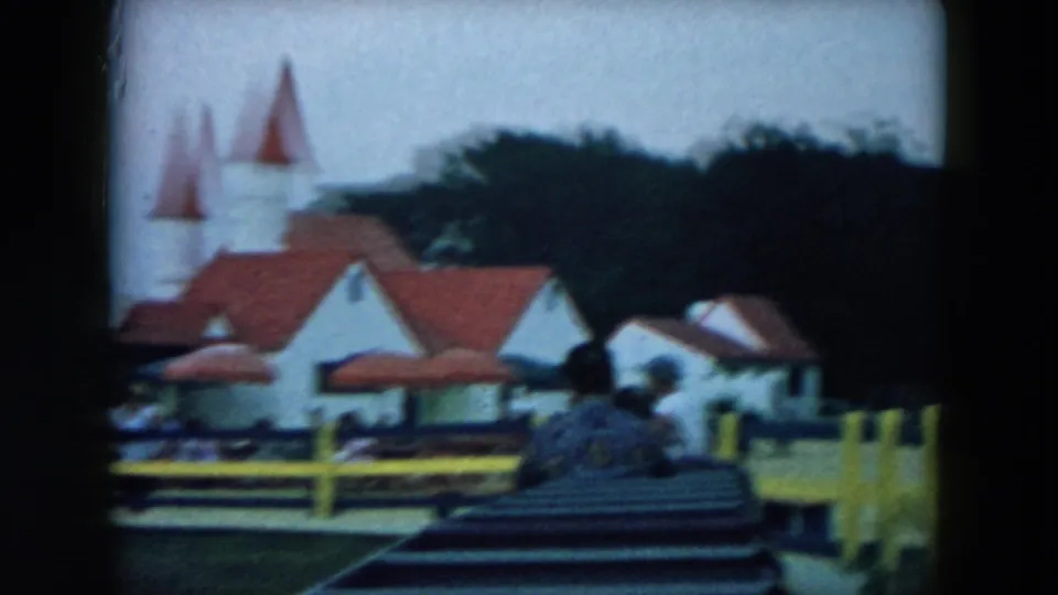 Home-movie ride view in Hicksville, New York, showing red-roofed white buildings, a seated visitor, yellow fencing, and trees.