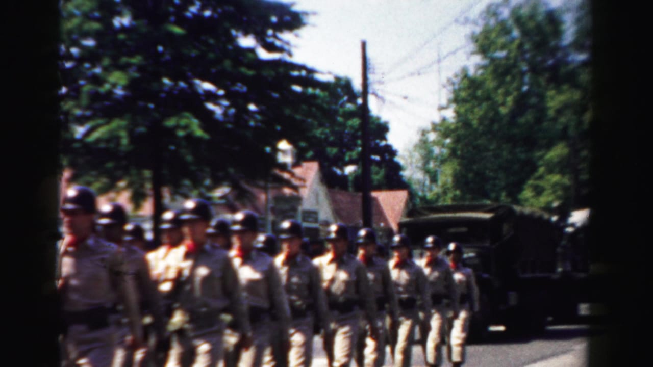 In Normal, Illinois in 1957, uniformed soldiers march through a town parade as a military truck follows nearby.