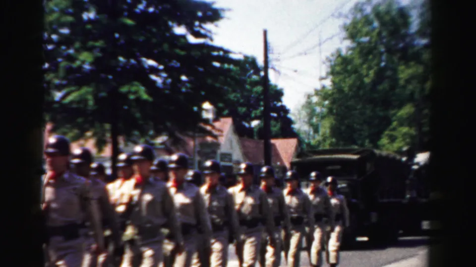 In Normal, Illinois in 1957, uniformed soldiers march through a town parade as a military truck follows nearby.