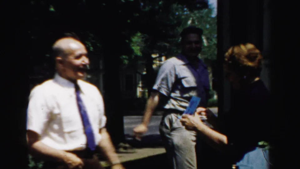 A smiling man in a white shirt and blue tie stands outside as nearby adults interact in shaded daylight.