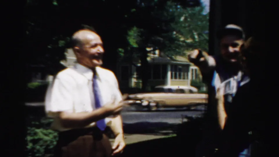 The smiling man remains in view near the house while another figure gestures from the shaded edge of the frame.