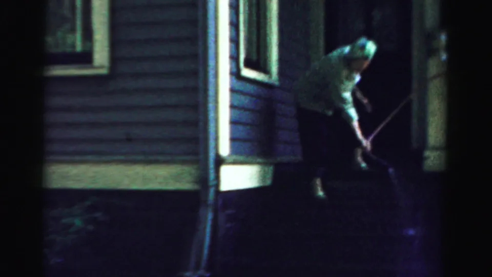 The older woman bends toward the porch steps with a hose beside the dark-sided house in the 1957 night scene.