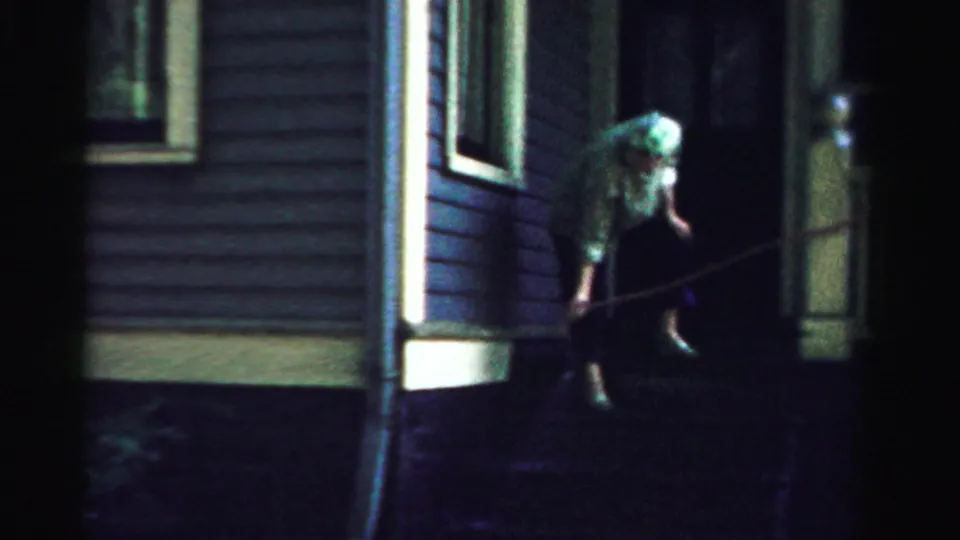 In the dim 1957 exterior, the woman continues bending over the front steps with a hose near the porch post.