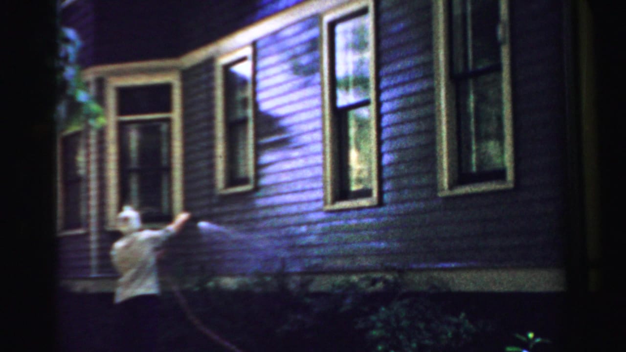 Older woman sprays the side of a painted house with a garden hose.