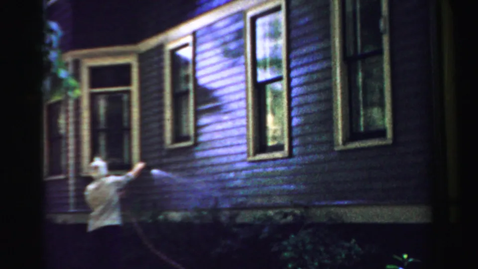 In Normal, Illinois, an older woman uses a garden hose on painted house siding beside tall windows.