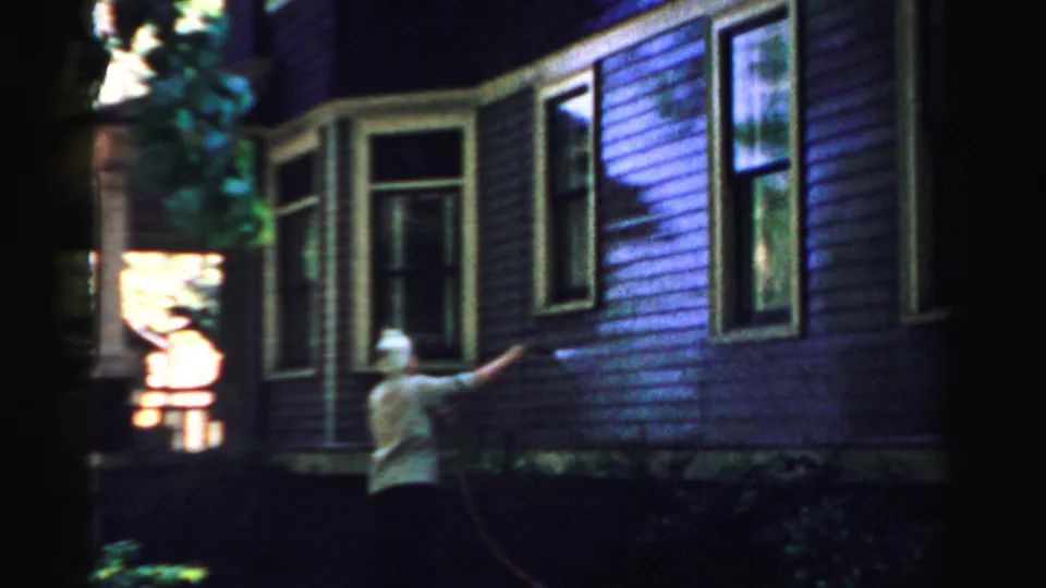 The woman continues spraying the house exterior, with porch trim and windows visible in the background.