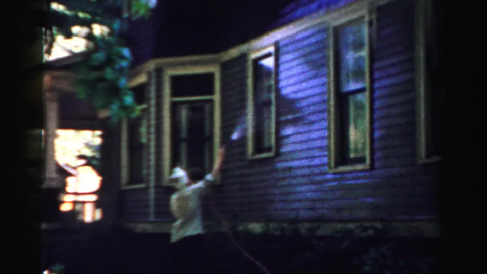 A 1957 home-movie frame shows the woman hosing the siding near a porch and tall window in Normal, Illinois.