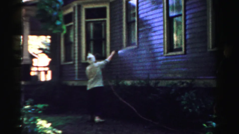 The woman raises the hose toward the upper siding, framed by windows, porch detail, and shaded plants.