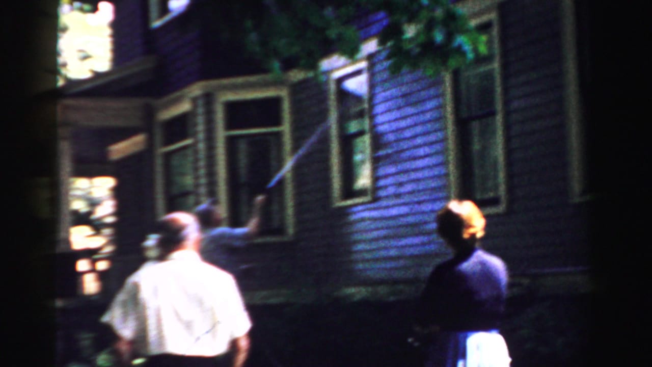 In Normal, Illinois, 1957 home-movie footage, a man sprays water at purple house siding as two adults stand nearby.
