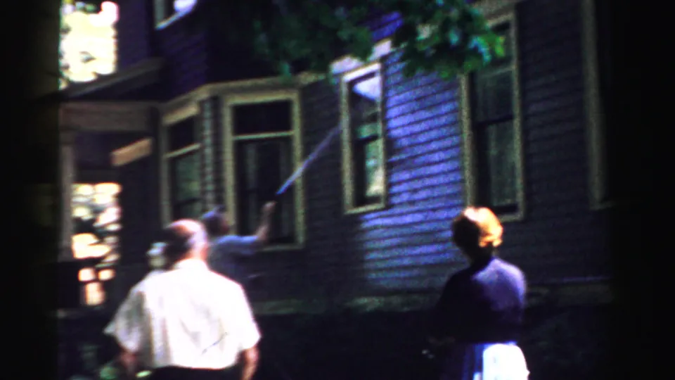 In Normal, Illinois, 1957 home-movie footage, a man sprays water at purple house siding as two adults stand nearby.