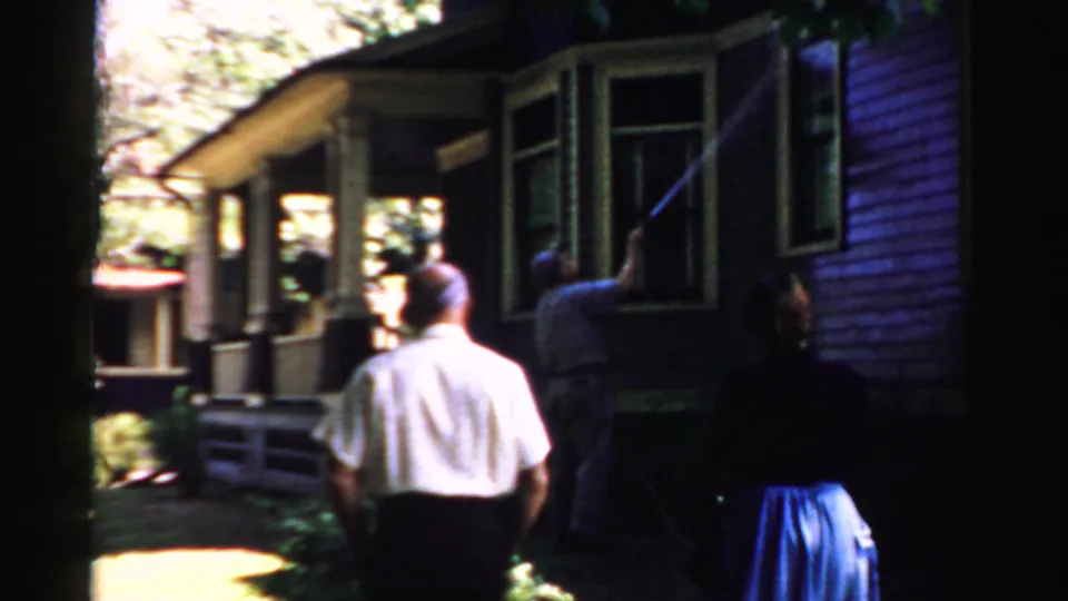 1957 Normal, Illinois footage shows a man washing a house window while two adult onlookers stand by the porch area.