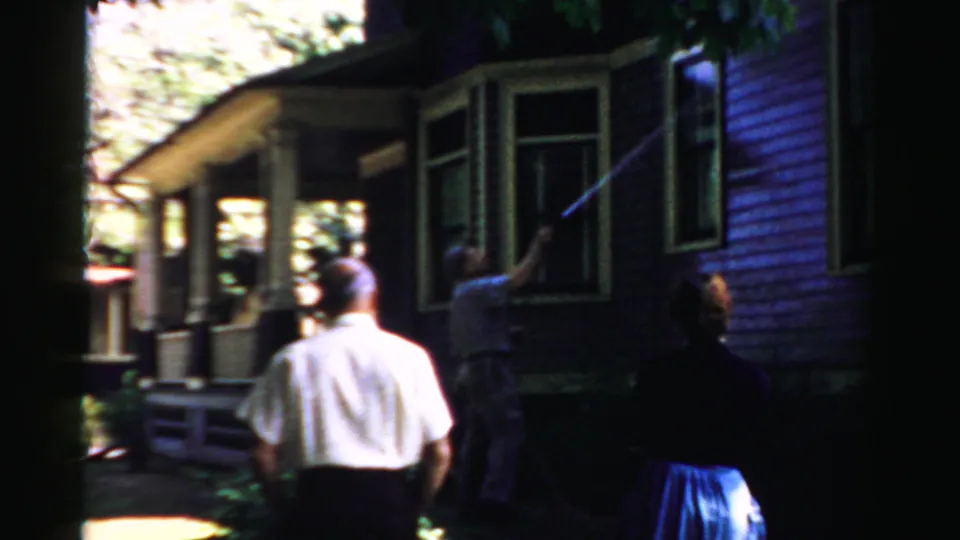 The home-movie frame shows an adult man power washing a window on a purple-sided house in Normal, Illinois, with observers nearby.