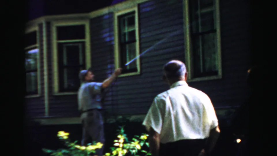 At the end of the clip, a man sprays the exterior siding and window area as an adult onlooker faces the house.