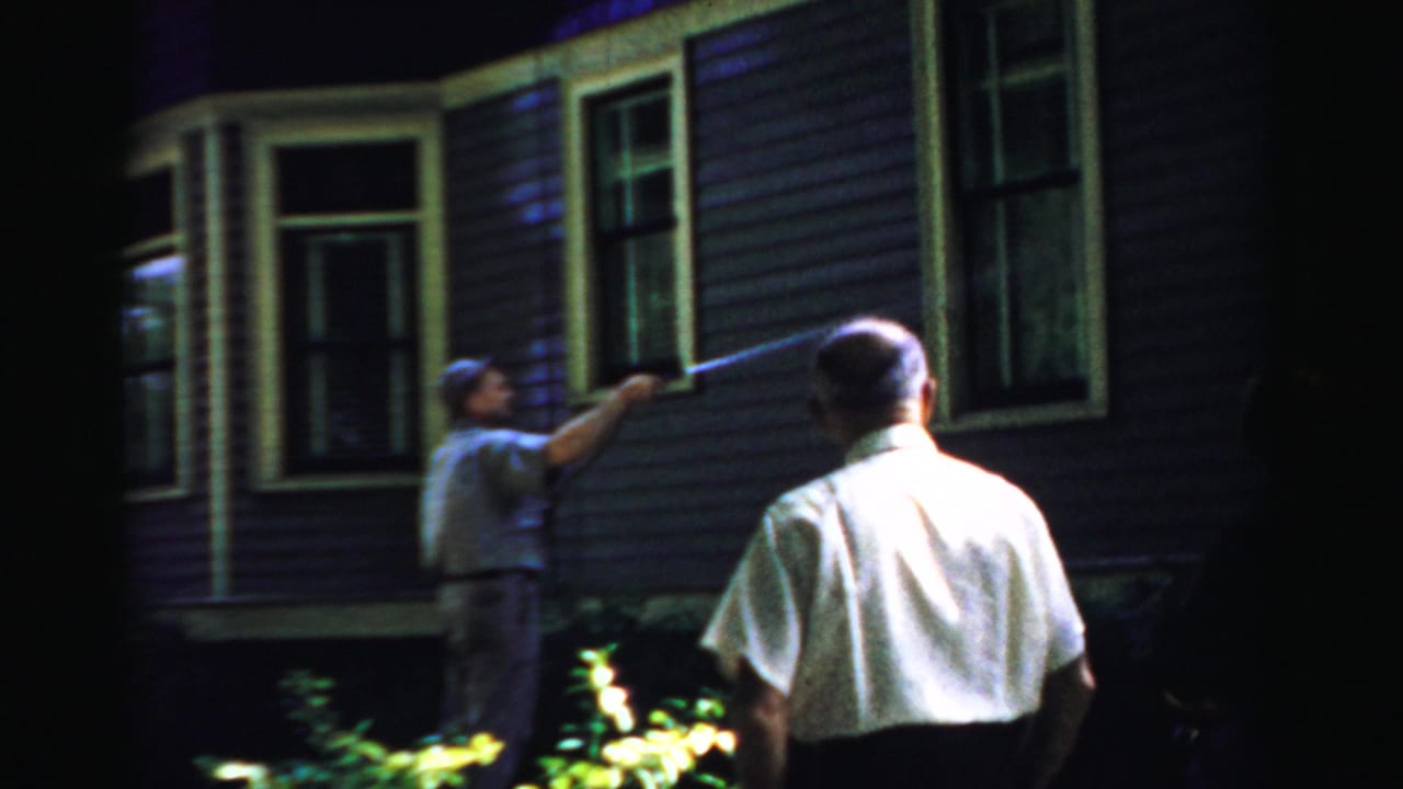 1957 Normal, Illinois 8mm frame showing a man using a hose on a house exterior at night as another man stands nearby.