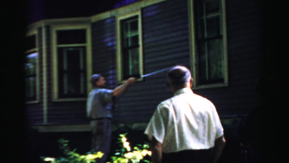 1957 Normal, Illinois 8mm frame showing a man using a hose on a house exterior at night as another man stands nearby.