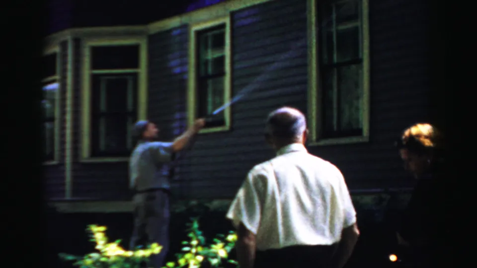 1957 Normal, Illinois home-movie frame of men outside a house at night, with one spraying water near a window.