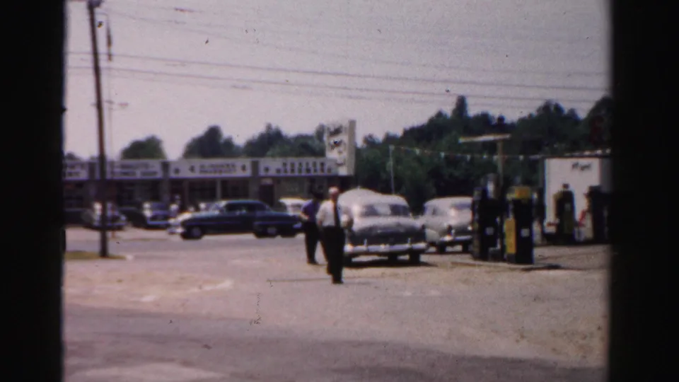 1957 Normal, Illinois 8mm frame showing men, mid-century cars, gas pumps, storefronts, and utility wires in daylight.